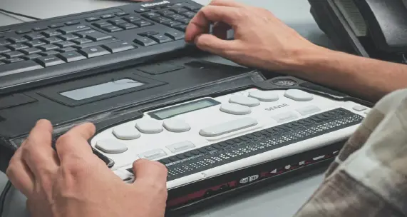 A man uses a braille display next to a computer keyboard on a desk.